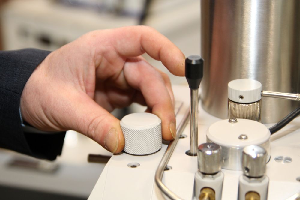 Zdjęcie przedstawia osobę obsługującą pokrętło na urządzeniu laboratoryjnym, prawdopodobnie w celu kontroli precyzyjnych parametrów