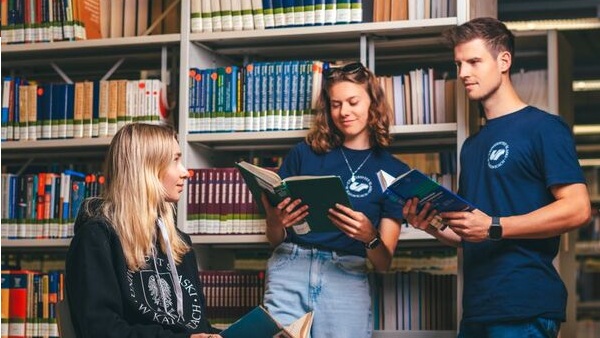 Trójka strudentów przy regale z ksiązkami, dwie dziewczyny i jeden chłopak. Osoby na zdjęciu prowadza ze sobą rozmowę.